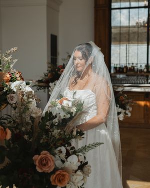 A bride in a flowing veil holds a romantic autumn-toned wedding bouquet with roses and eucalyptus in a rustic venue.