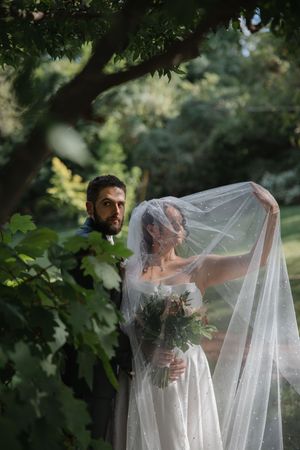 A romantic wedding photo through tree branches shows a bride in a flowing veil holding a bouquet in a garden setting.