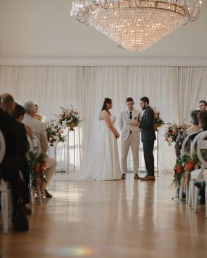 A wedding ceremony takes place in an elegant white ballroom with crystal chandeliers and draped fabric decor.