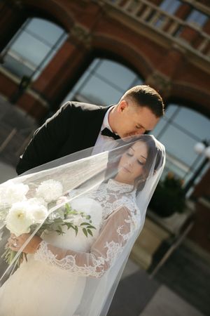 A romantic bride and groom share an intimate moment under flowing white veil against a brick building background.