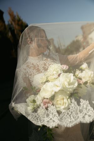 A wedding veil with delicate lace trim drapes over white roses and floral arrangements in soft natural lighting.