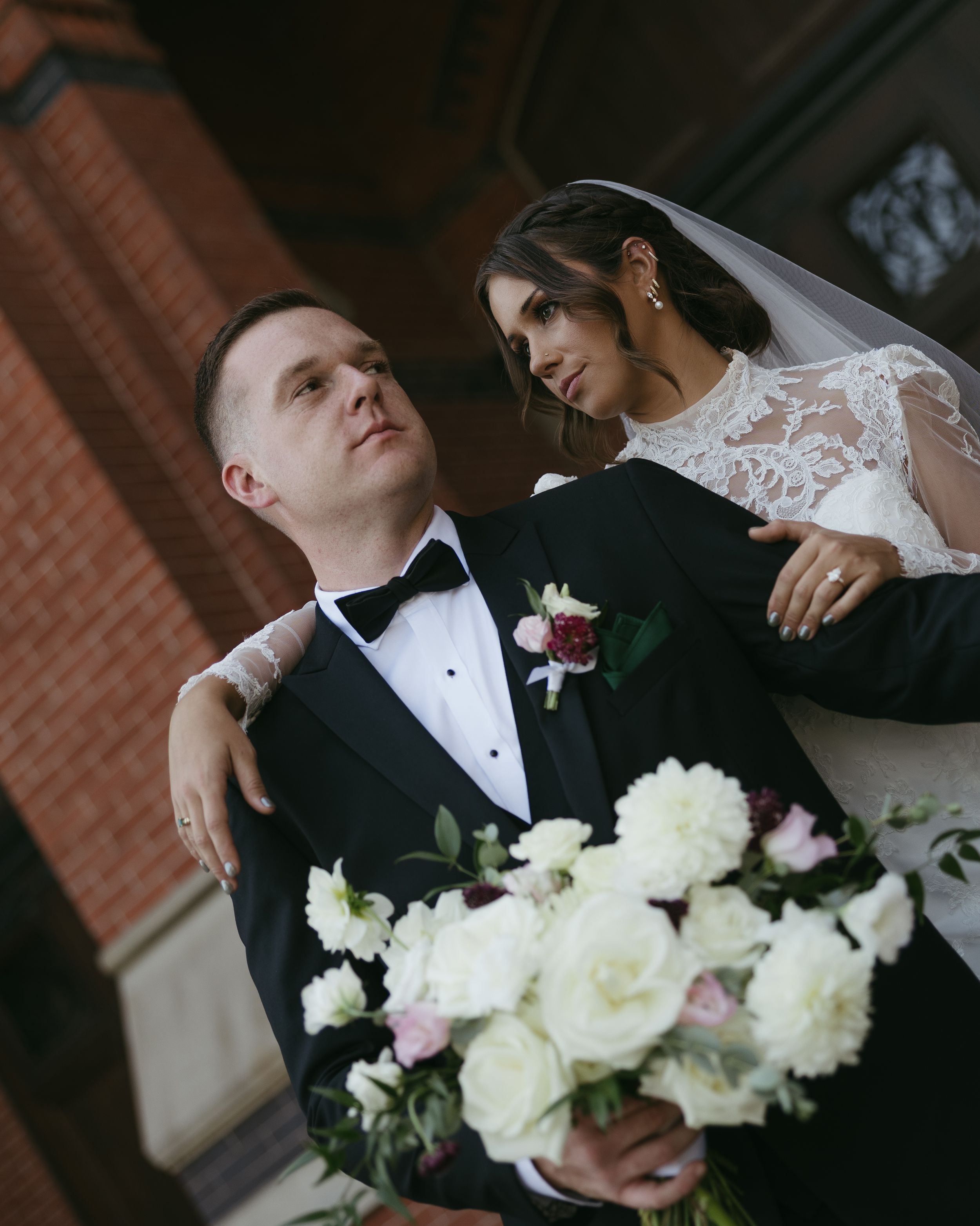Newlywed couple poses with beautiful white and pink roses against brick building backdrop in elegant wedding attire.