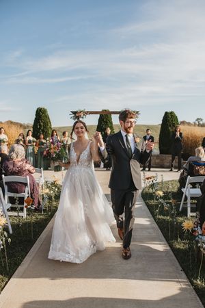 Wedding ceremony processional at outdoor venue with stone walls and white chairs on concrete path under blue sky.