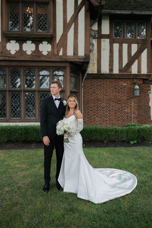 A wedding couple poses in front of a Tudor-style building with ornate windows and brick details.