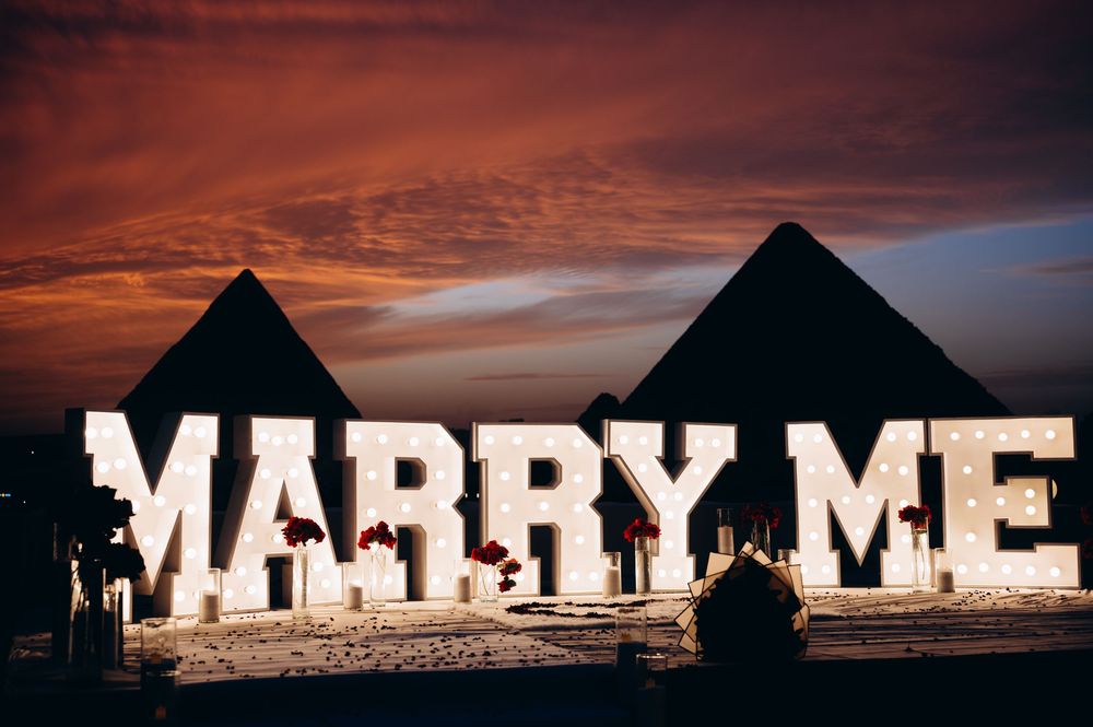Giant illuminated MARRY ME letters spell out a proposal at sunset against a dramatic sky.