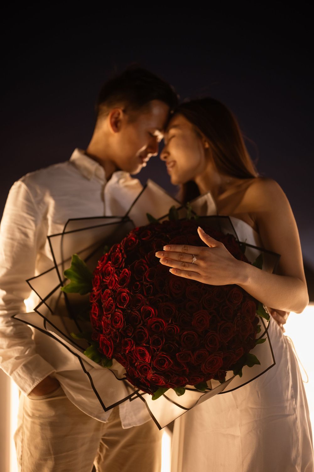 A romantic couple in white clothing share intimate moments while holding a bouquet of red roses against a dark background.