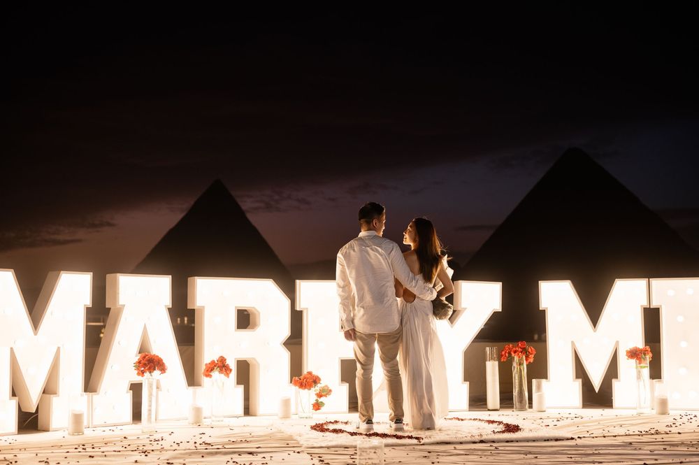 Large lighted letters spelling MARRY ME line a beach at dusk.