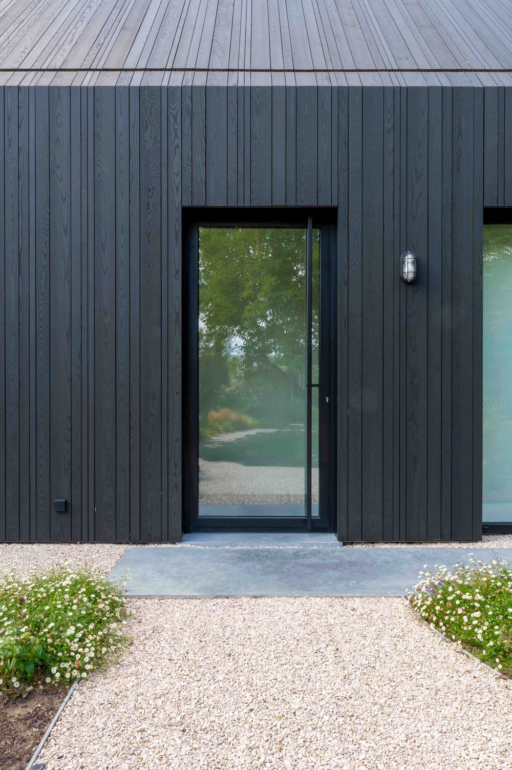 Modern black wooden building exterior with white gravel path and small flowering plants along the entrance.