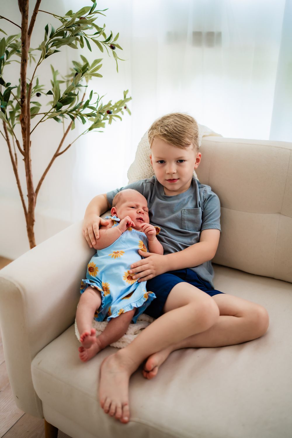 Two siblings share a tender moment together on a beige couch with potted plants in the background.
