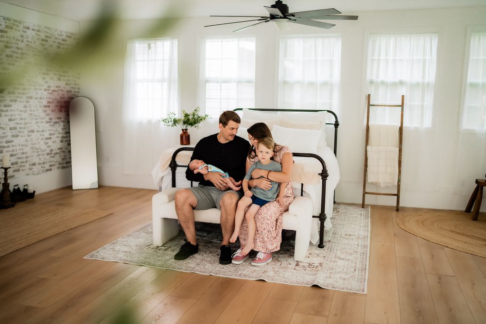 A couple sits on a hardwood floor in a bright, minimalist interior space with white walls and sheer curtains.