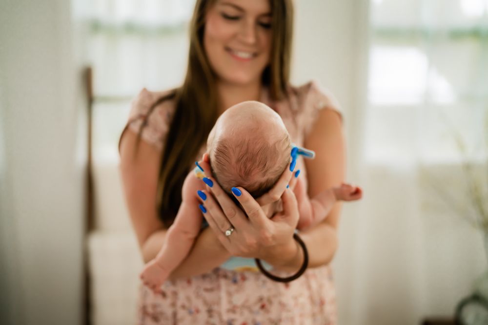 A happy parent in a floral dress holds and gazes lovingly at a newborn baby inside a bright, sunlit room.