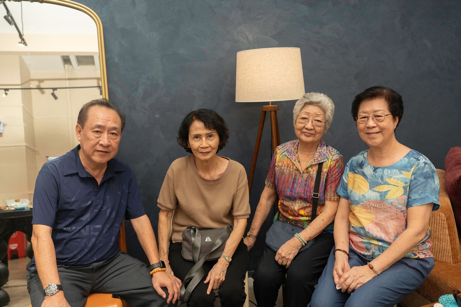 A small group sits together smiling on a sofa in front of a gray wall with a standing lamp in the background.
