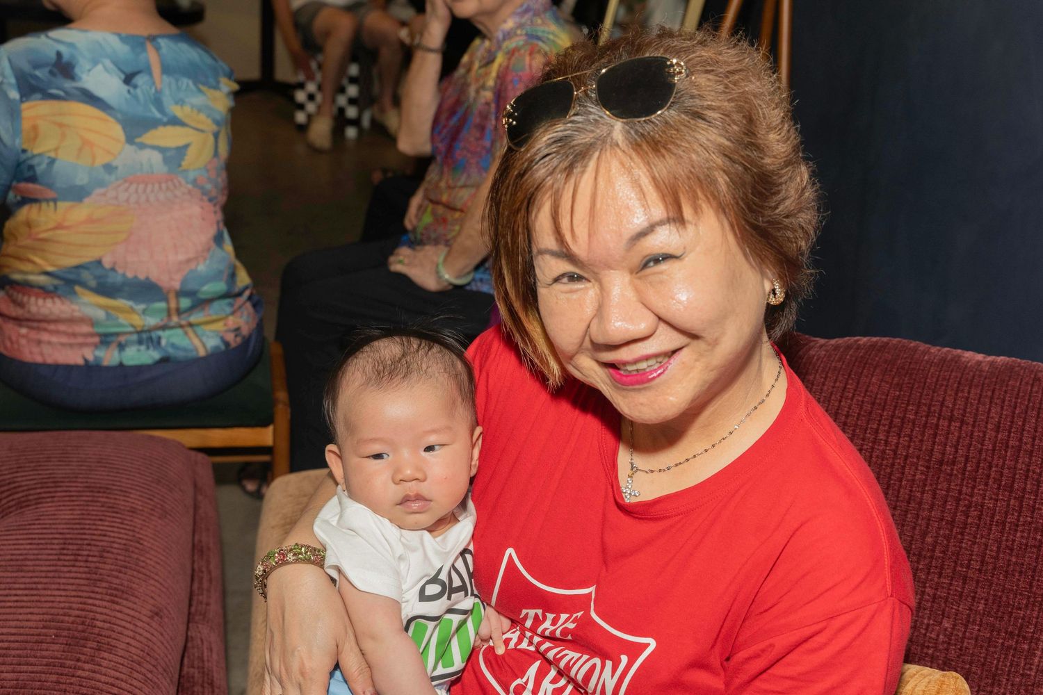 A woman in a red t-shirt sits on a brown couch holding a young baby wearing a white outfit.