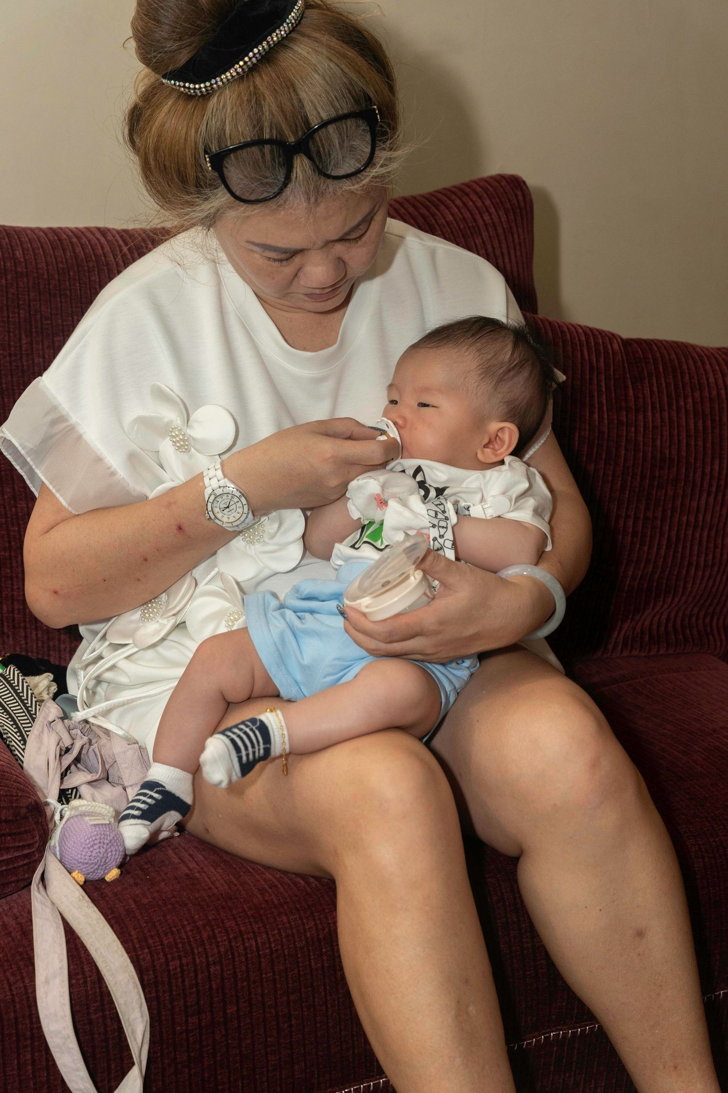 An adult sitting on a burgundy couch bottle feeds a young baby wearing blue and white clothes.