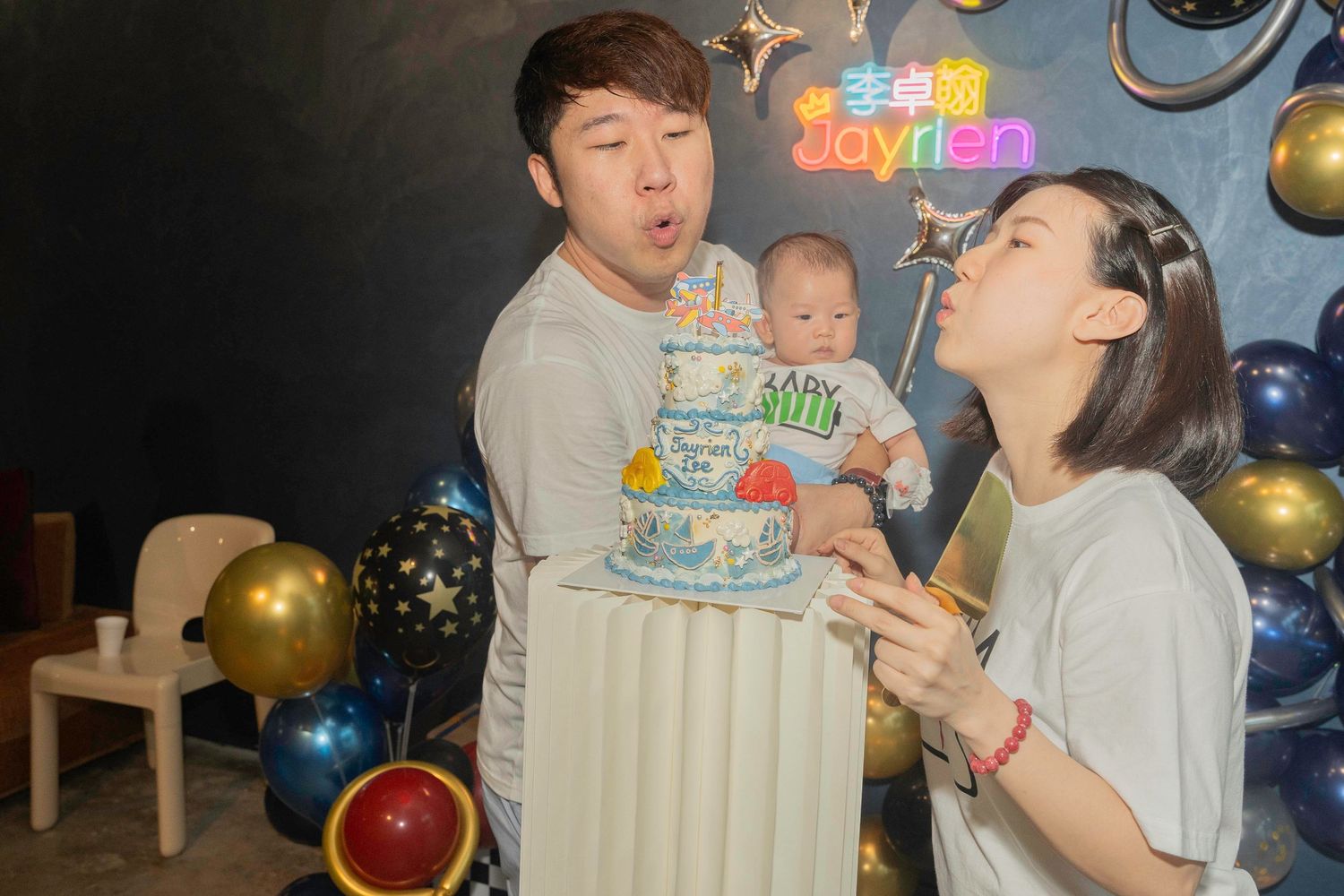 A birthday celebration with balloons and family members viewing a decorated cake with blue frosting at home.