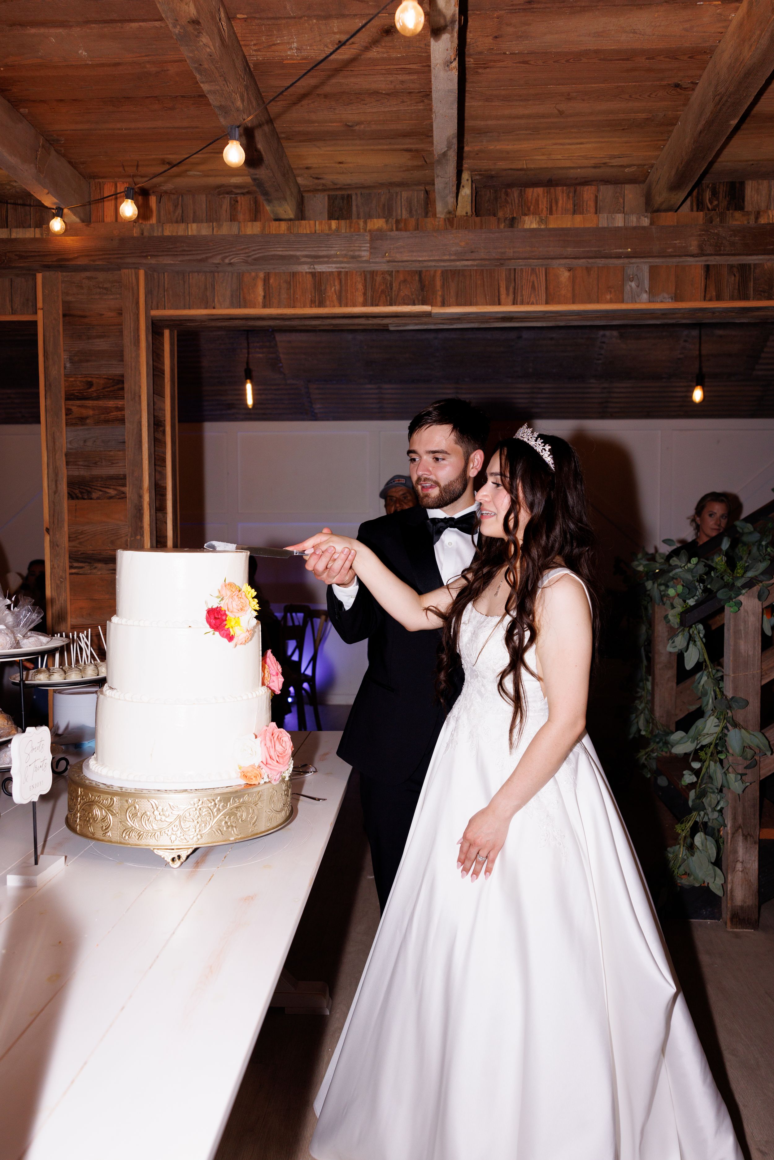 Wedding couple cuts their elegant white and gold cake in rustic wooden reception venue.