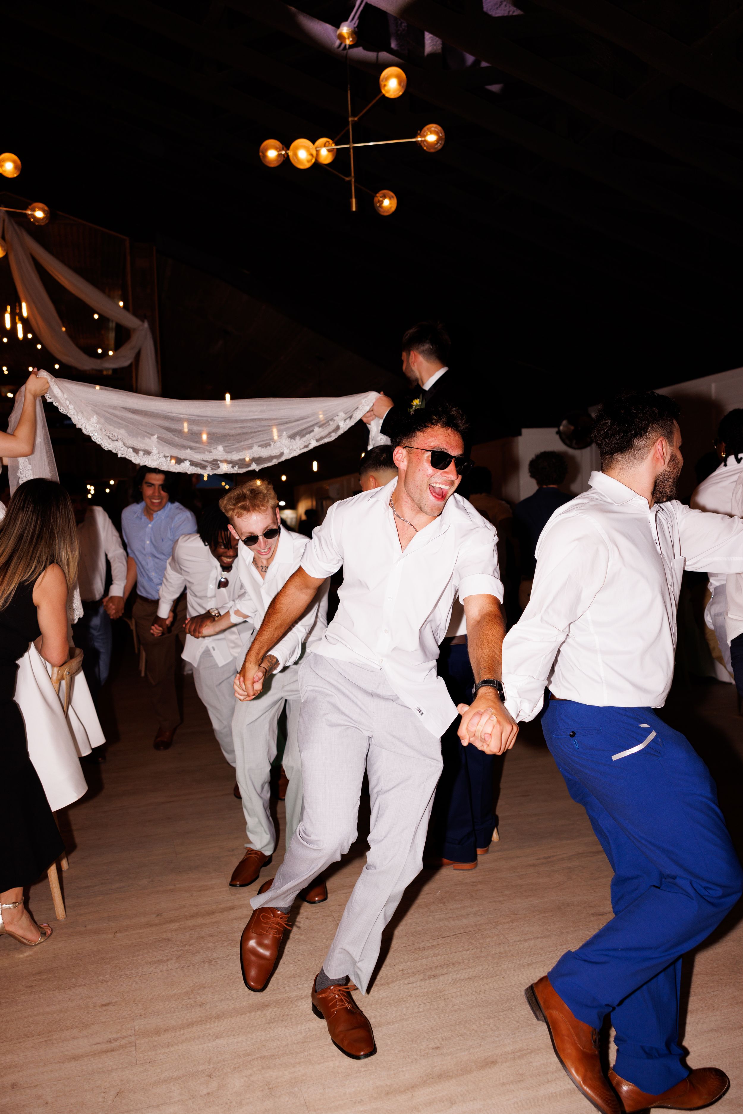 People in white outfits perform energetic dance moves at a wedding reception.