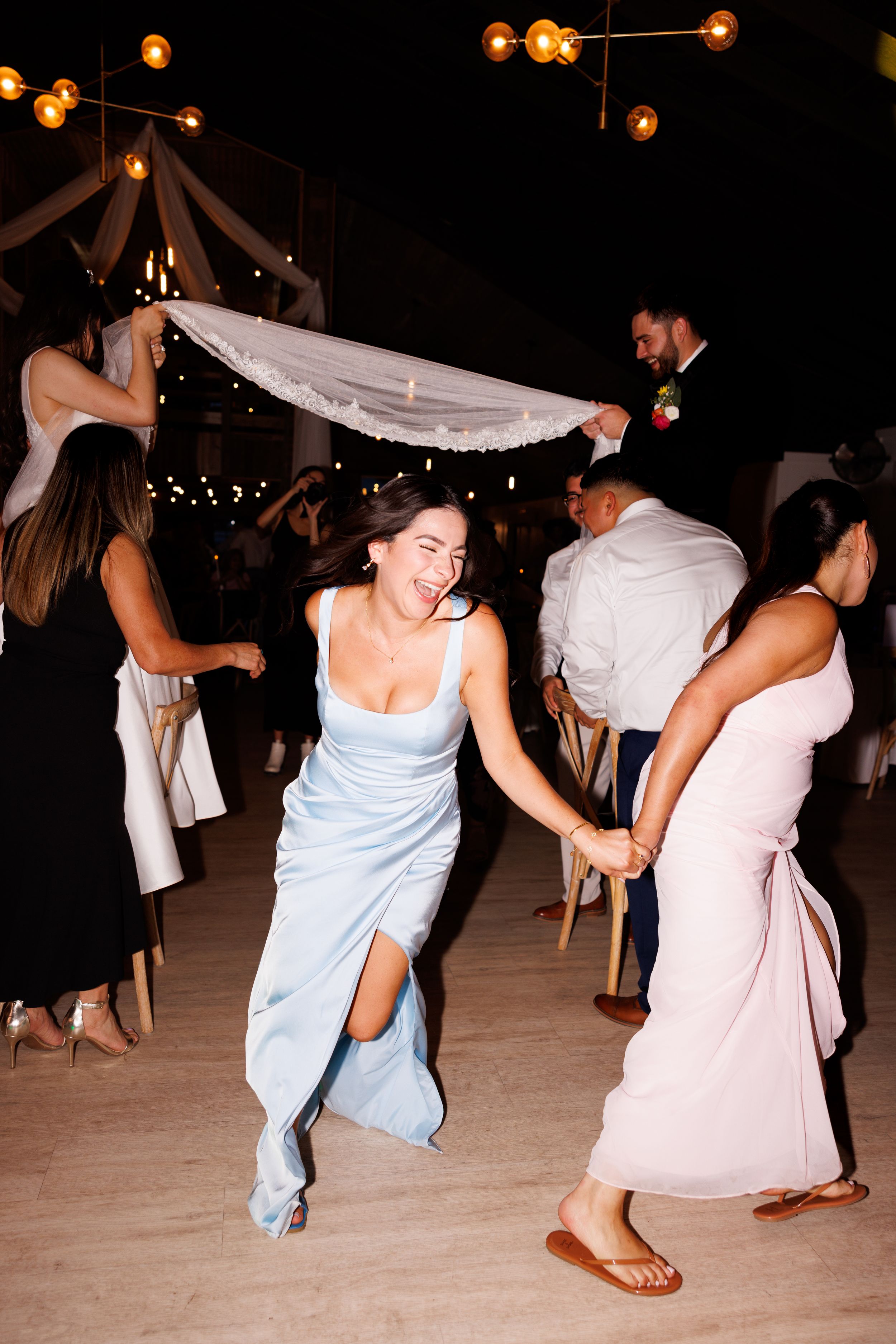 Guests in formal attire dance enthusiastically on a wooden floor under hanging lights.