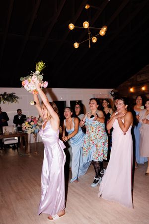 Women in light-colored dresses dance with bouquets at a wedding reception.