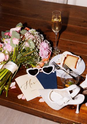 Wedding accessories including pink flowers, white sunglasses, and shoes arranged on a wooden table.