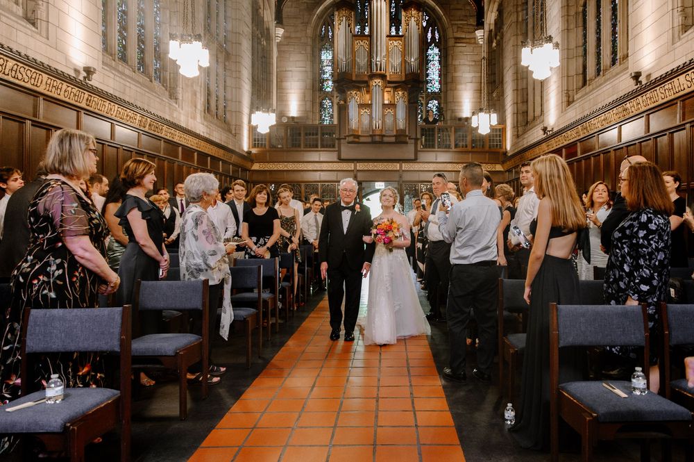 Group of elegantly dressed wedding guests mingling in a grand ballroom with ornate architectural details and warm lighting.