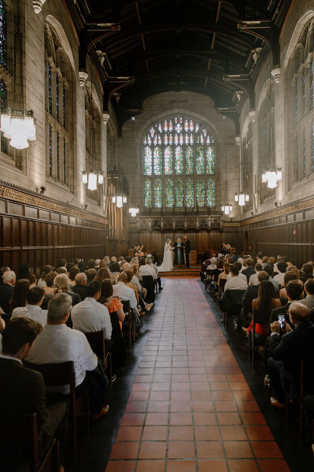 A wedding ceremony in a grand Gothic-style chapel with wooden pews and a stunning stained glass window.