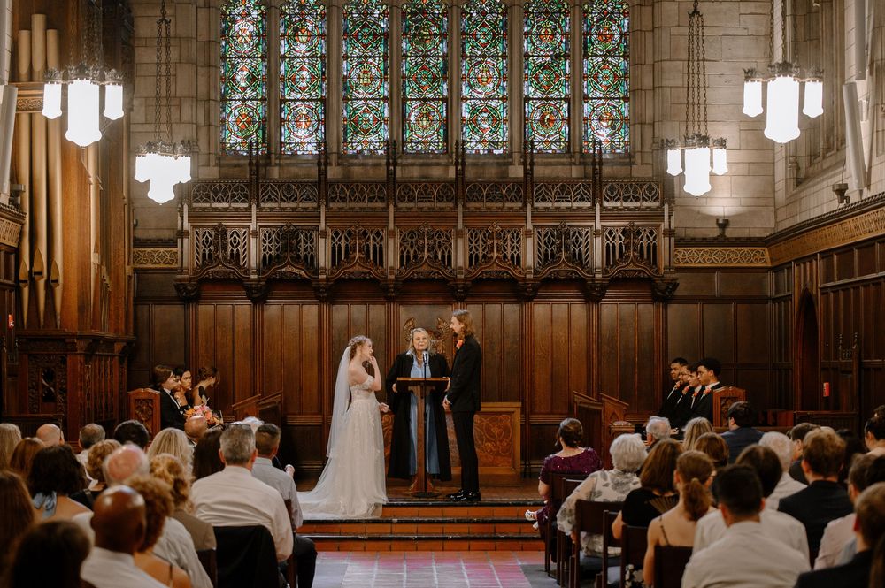 A wedding ceremony takes place inside a historic chapel with wooden paneling and stained glass windows.