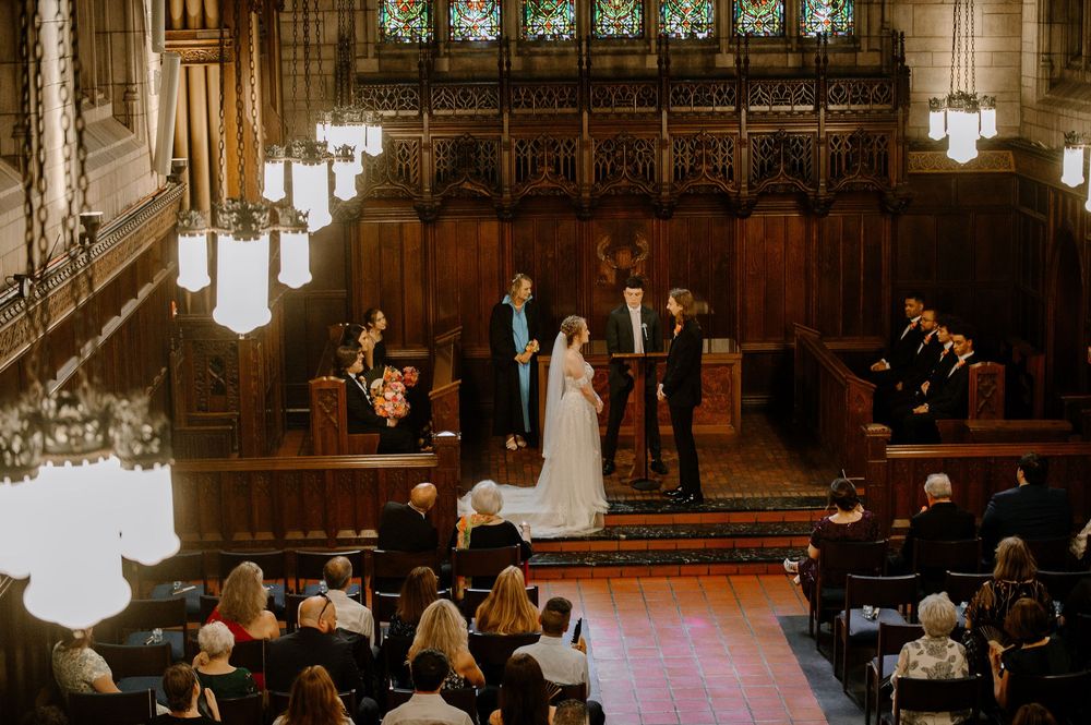 A wedding ceremony takes place inside a grand historic chapel with ornate wood paneling and glowing pendant lights.