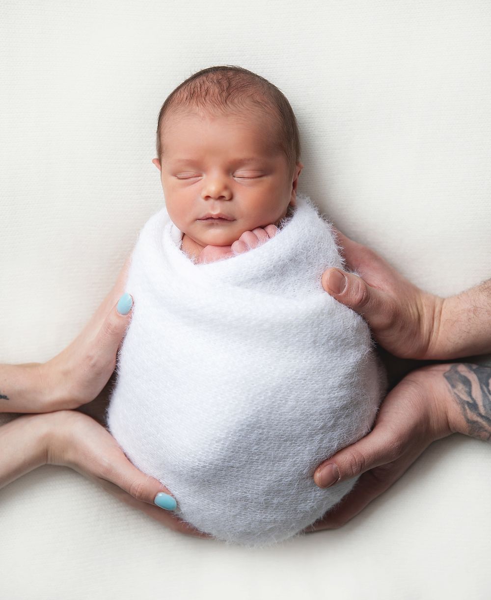 A sleeping newborn baby swaddled in white cloth is gently cradled in supportive hands. During a newborn session in Sidcup