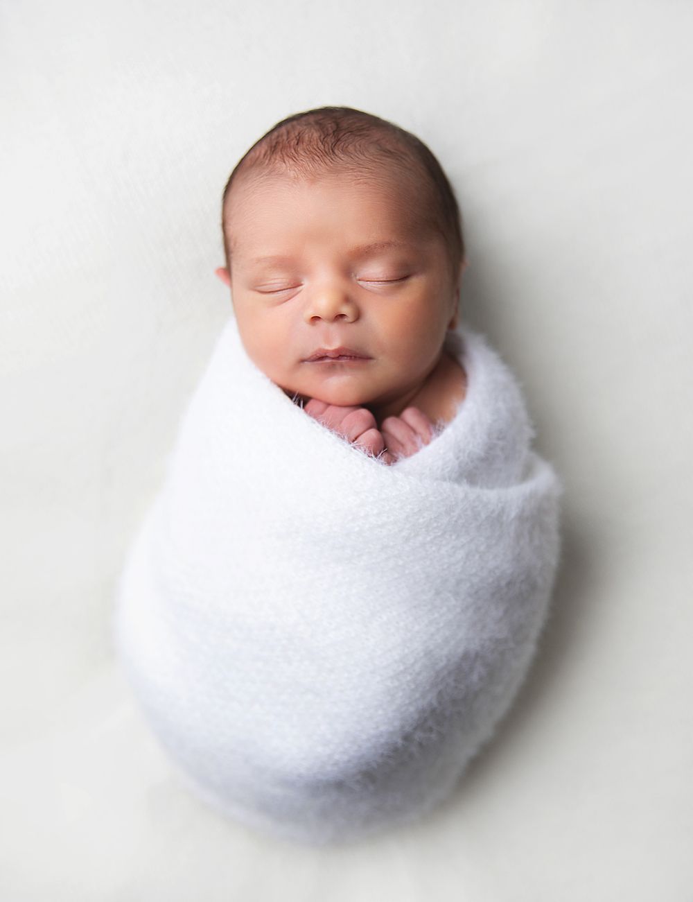 Sleeping newborn baby peacefully wrapped in soft white blanket against light background. During a newborn photoshoot in Sidcup