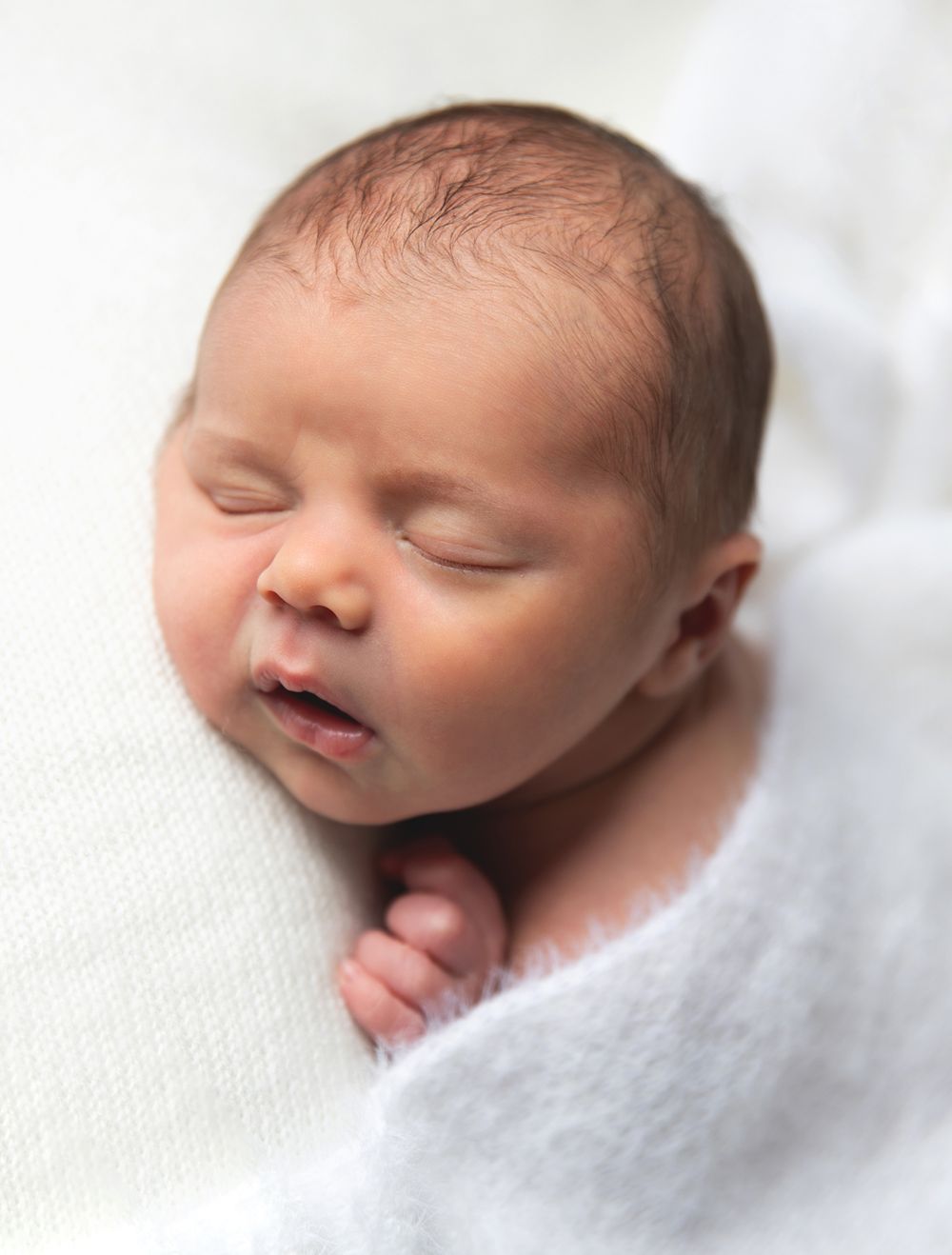 A peaceful sleeping newborn baby swaddled in soft white blankets against a light background. During a newborn photoshoot in Sidcup