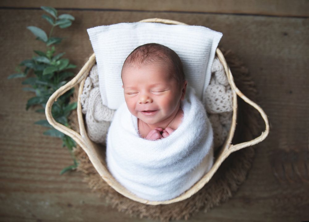 Newborn resting in rustic wooden basket lined with white fabric against woodgrain background. During a newborn photoshoot in Sidcup
