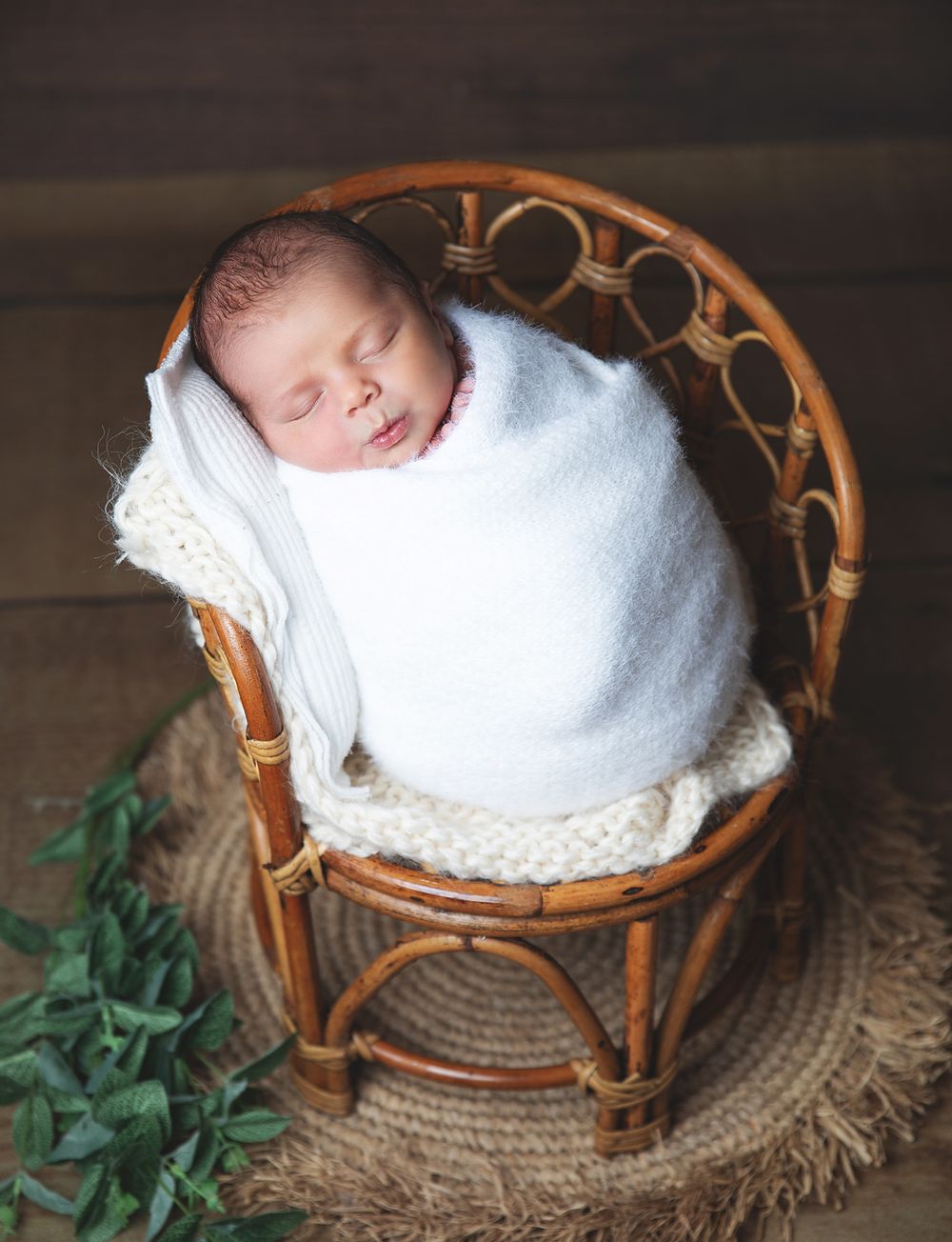 Sleeping newborn baby swaddled in white blanket resting in wicker chair with decorative greenery. During a newborn photoshoot in Sidcup
