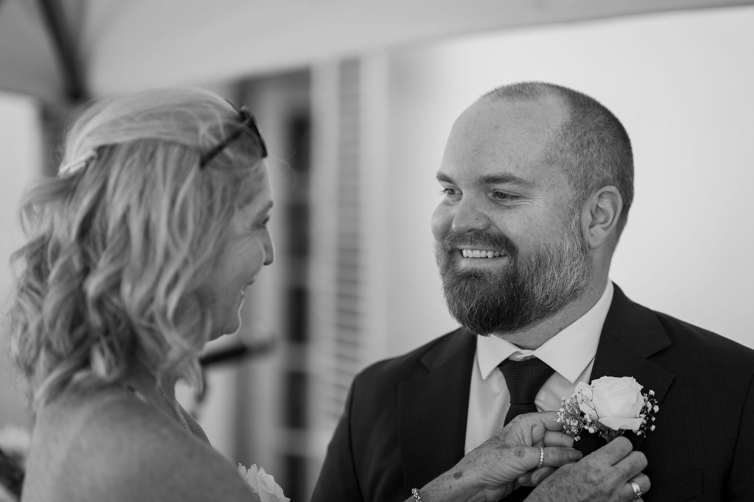An elderly person in a tan suit and striped tie smiles as someone pins a white boutonniere on their lapel.