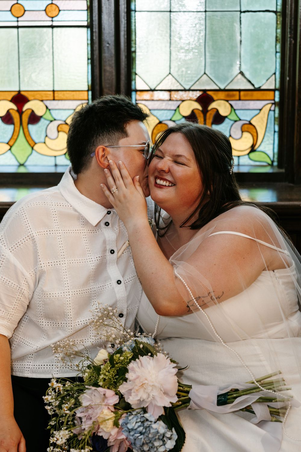 Bride laughing joyfully as she holds groom's face near stained glass window, holding pink floral bouquet.