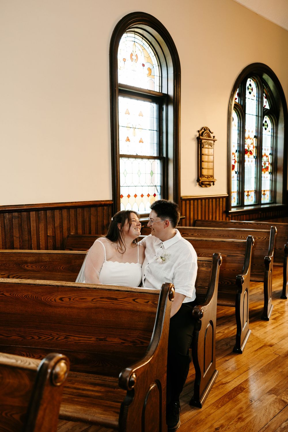 Couple sharing intimate moment on wooden church pew with arched stained glass windows in background.