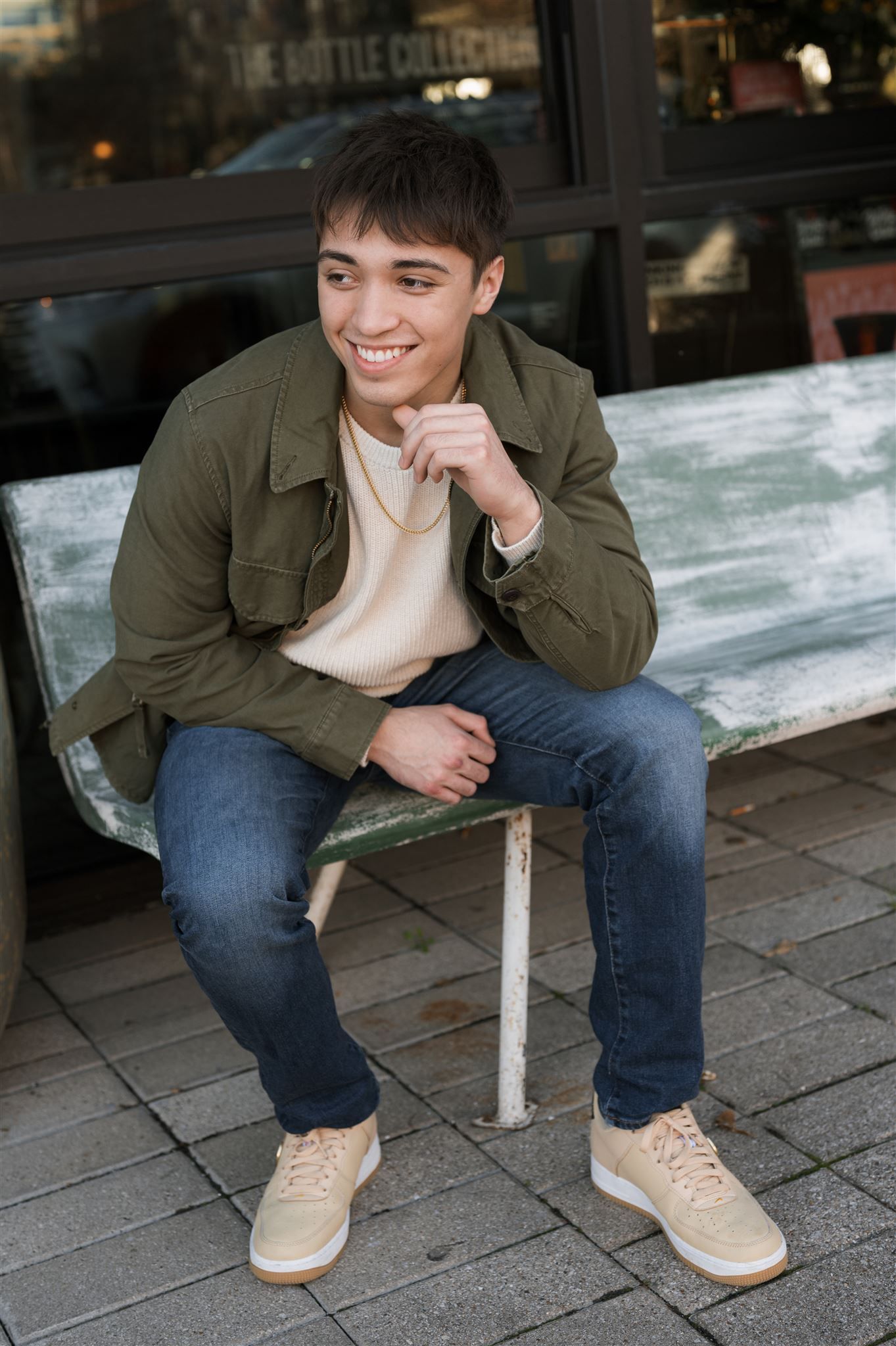 High school senior guy in olive jacket and jeans sits on bench wearing tan suede shoes creating casual portrait series.