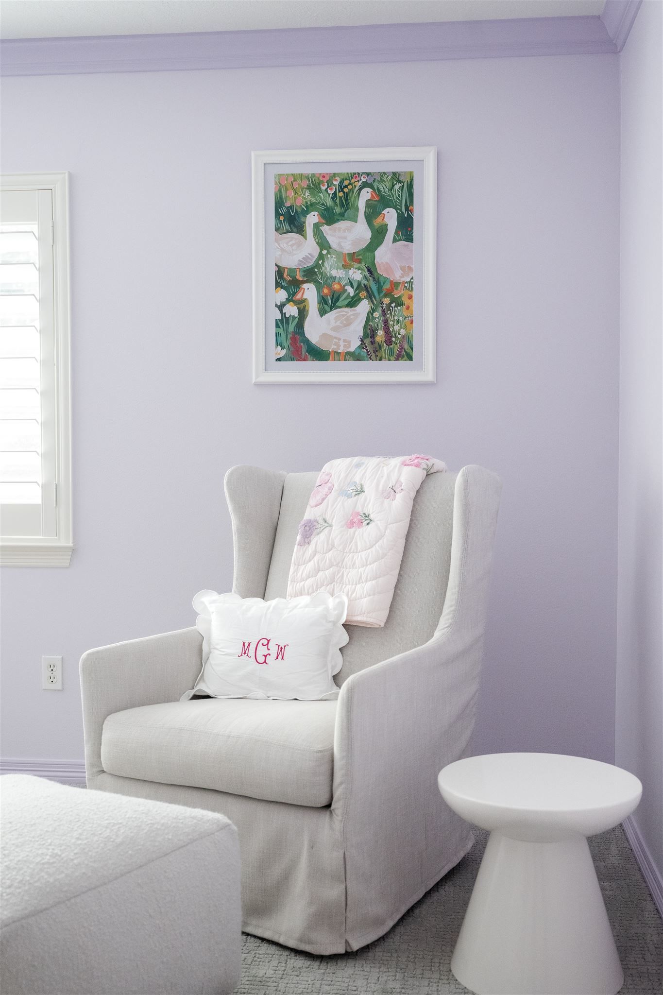 A cozy nursery corner with a light gray wingback chair, decorative goose artwork, and lavender walls.
