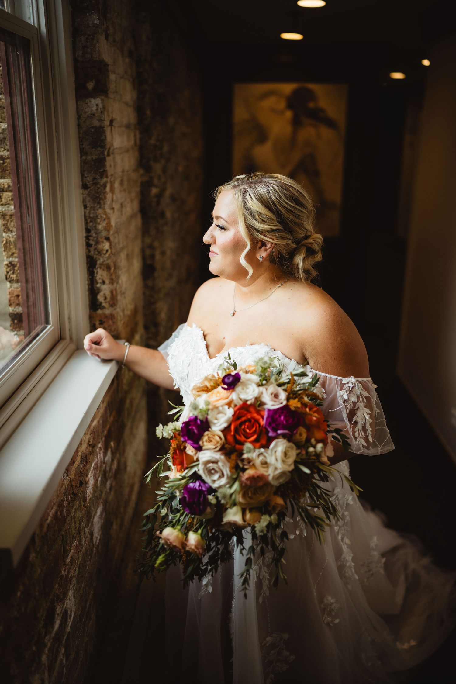 A bride in a flowing white wedding gown stands beside a brick building with ivy-covered walls.