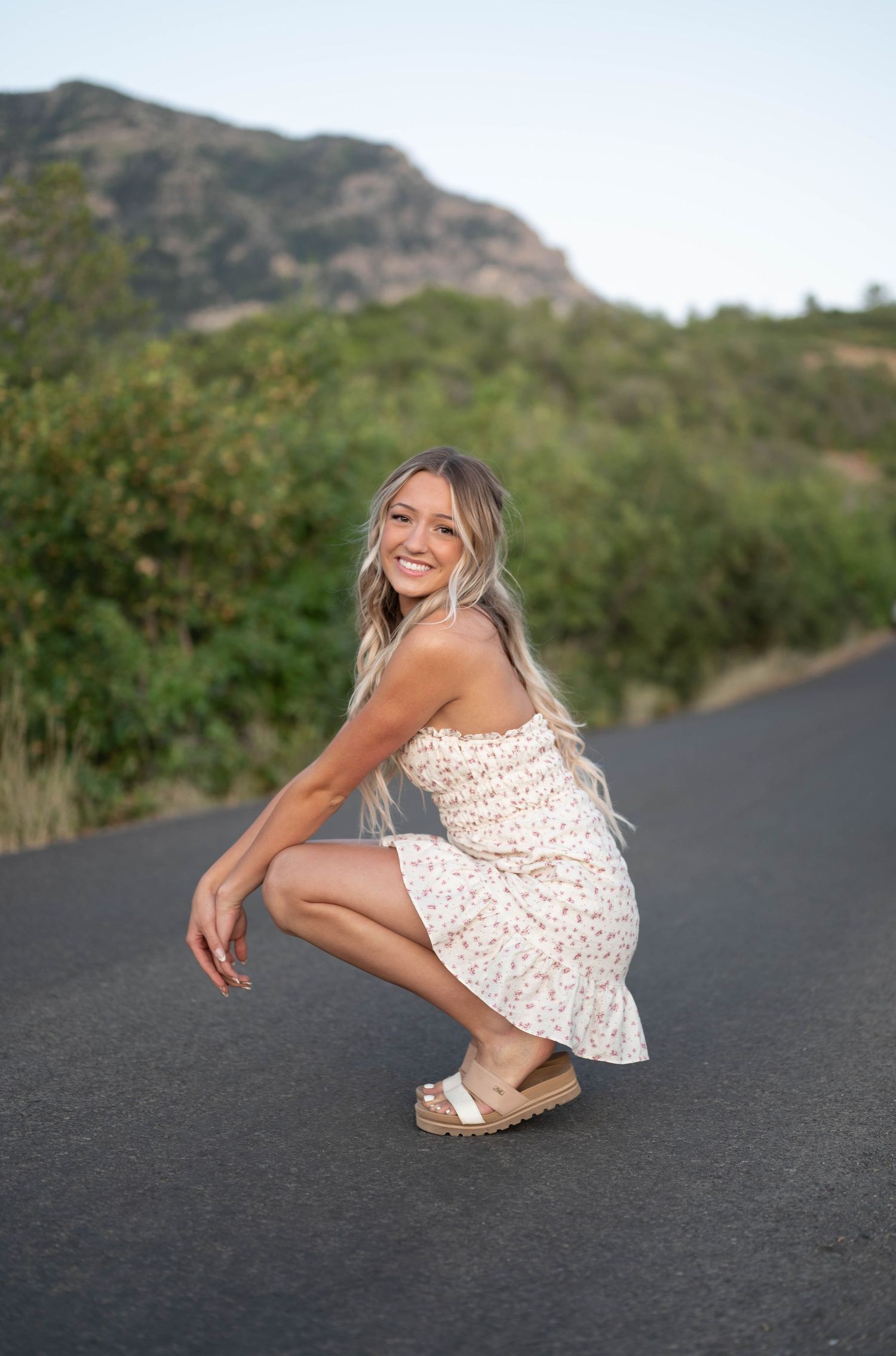 Person in white floral dress poses casually on an asphalt road with mountains in background.