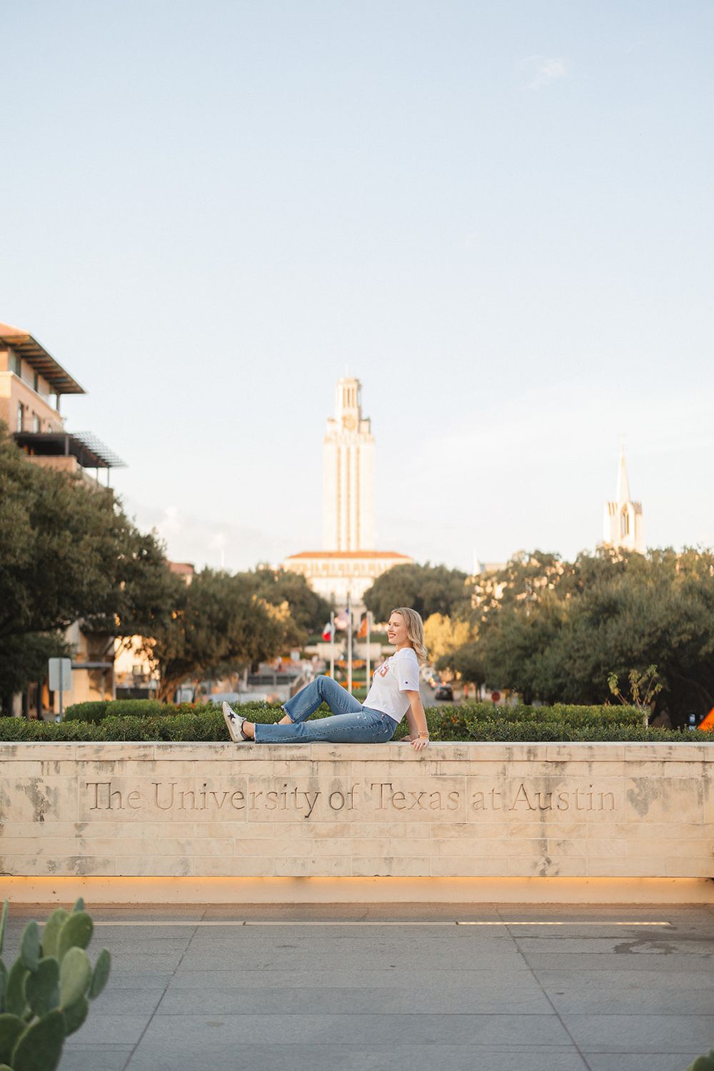 Iconic University of Texas tower glows orange in the sunset behind stone walls and cacti on campus.
