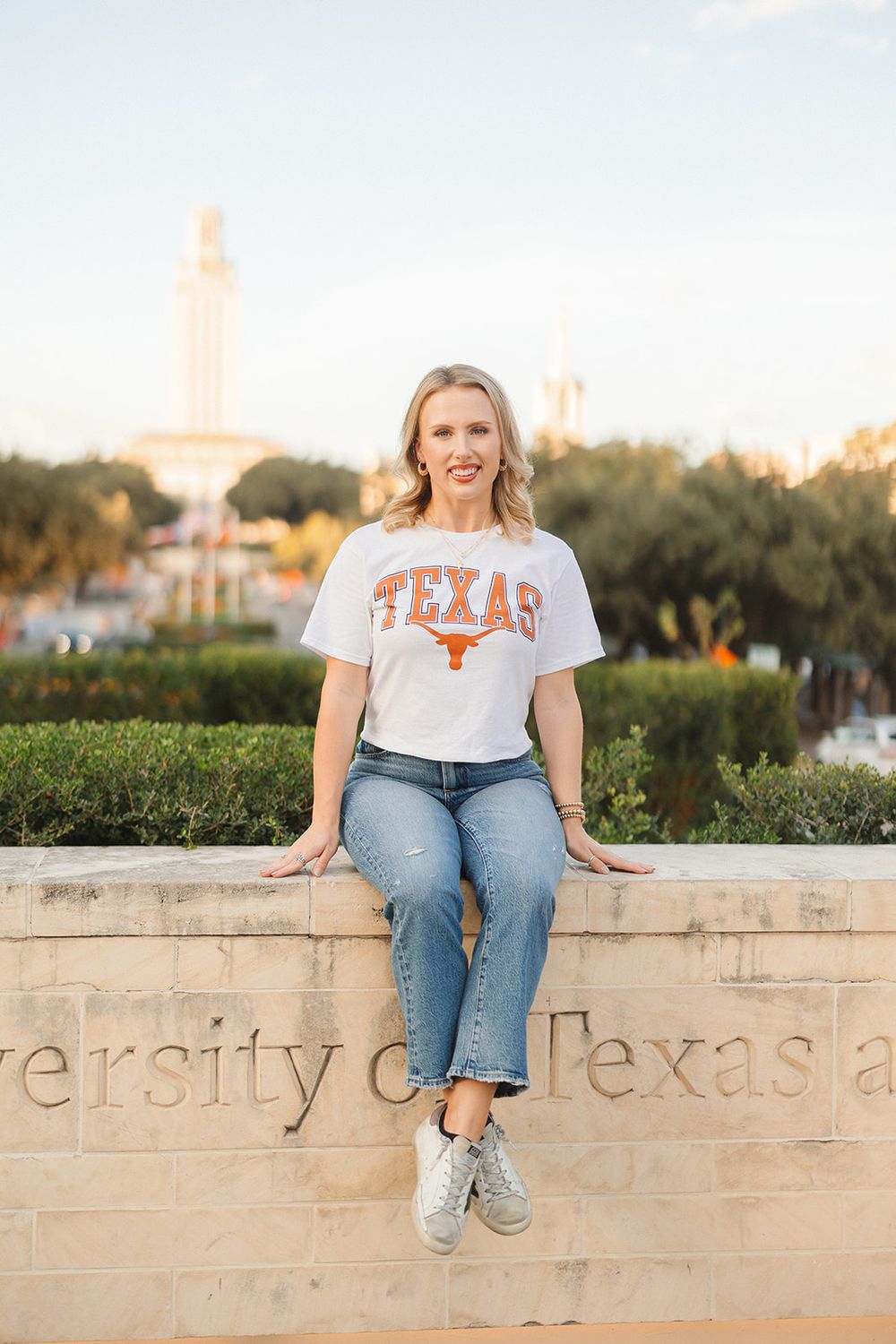 Student sits on University of Texas stone wall wearing branded t-shirt with iconic UT Tower visible in background.