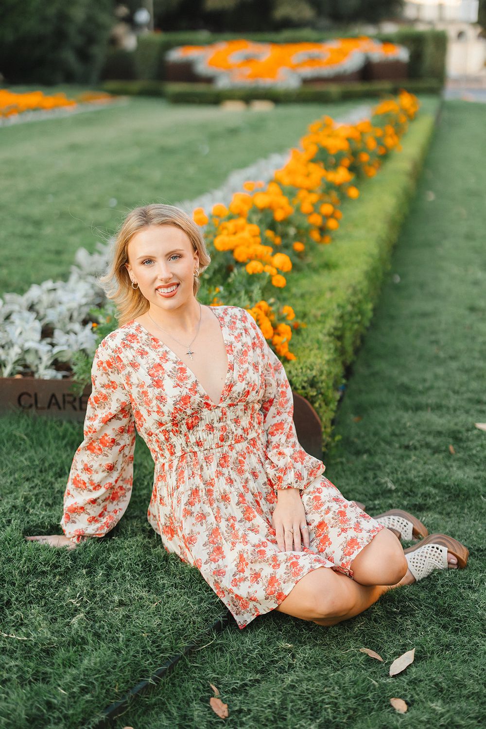 Person in a floral print dress poses on manicured lawn with orange marigold flowers in garden background.