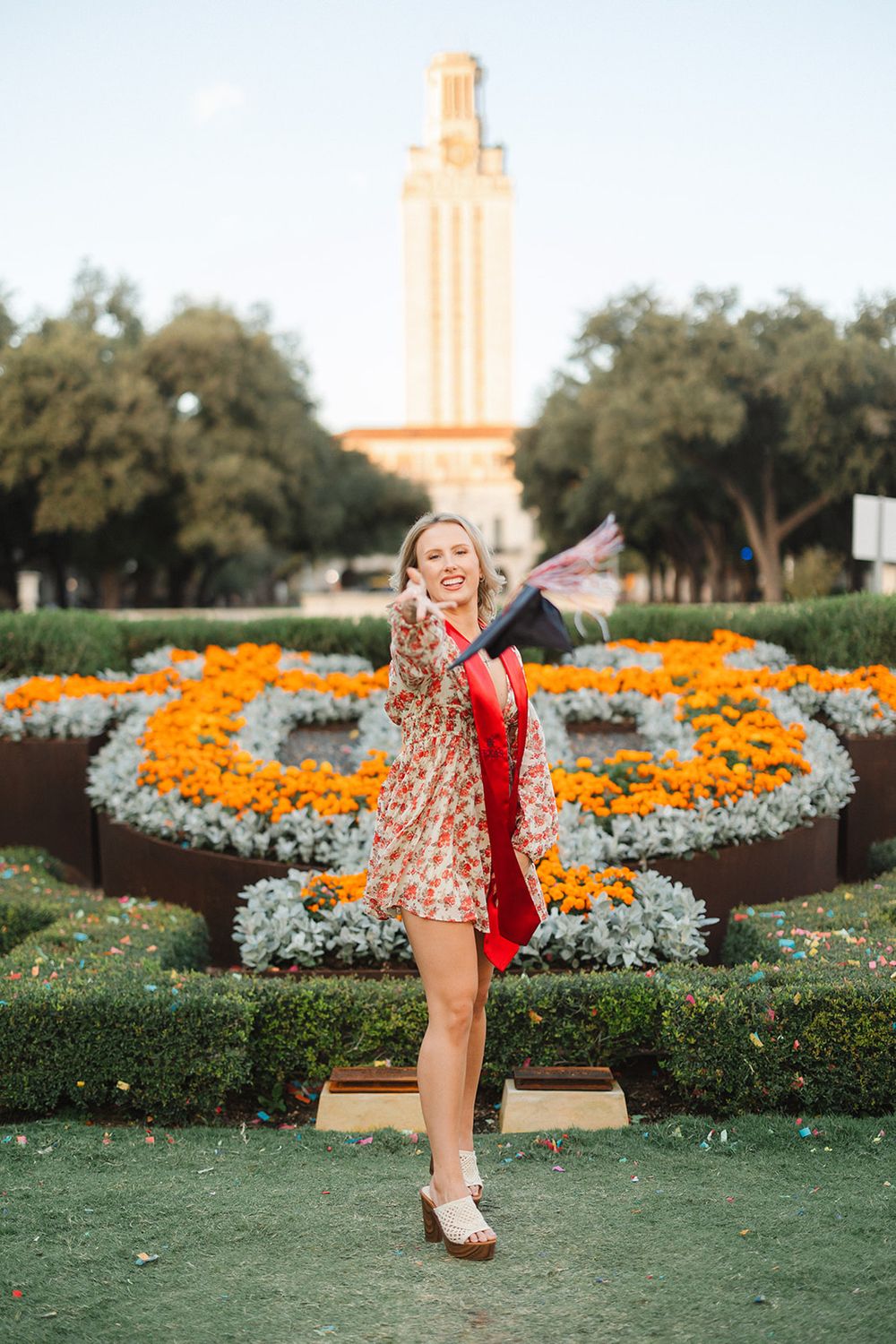 Graduate poses joyfully in floral dress with red stole at UT Austin campus with iconic tower and flower garden in background.