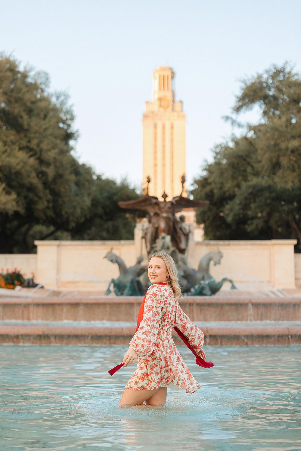 A person wades in a fountain pool wearing a floral dress with the UT Austin Tower visible in the background at sunset.