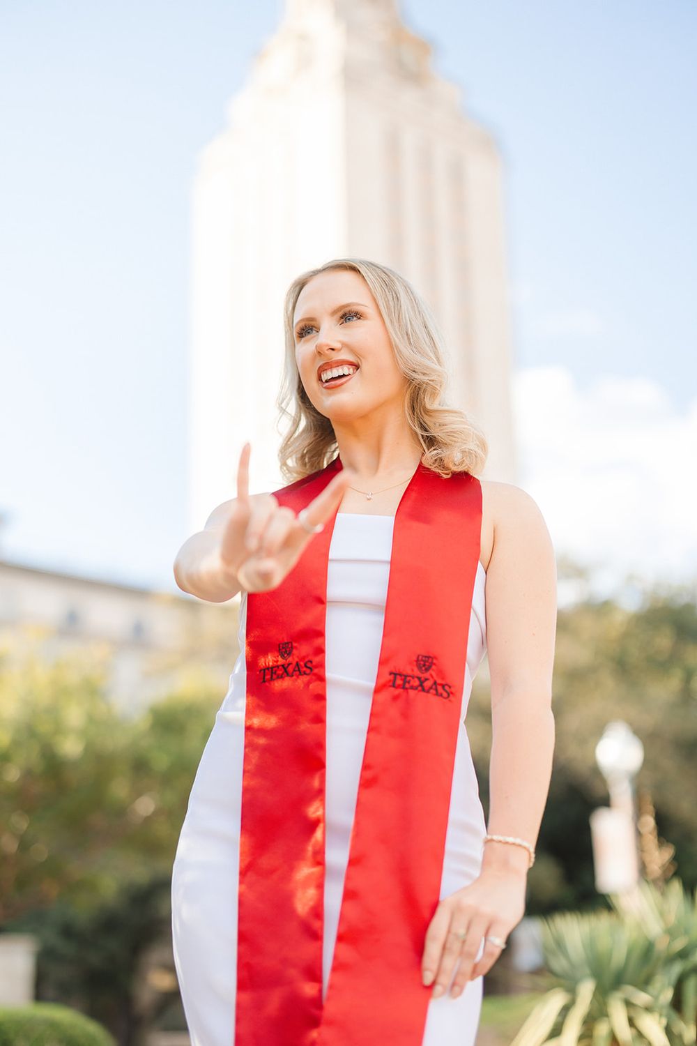 Graduate in white dress and red graduation stole smiles joyfully in front of sunlit tower on campus.