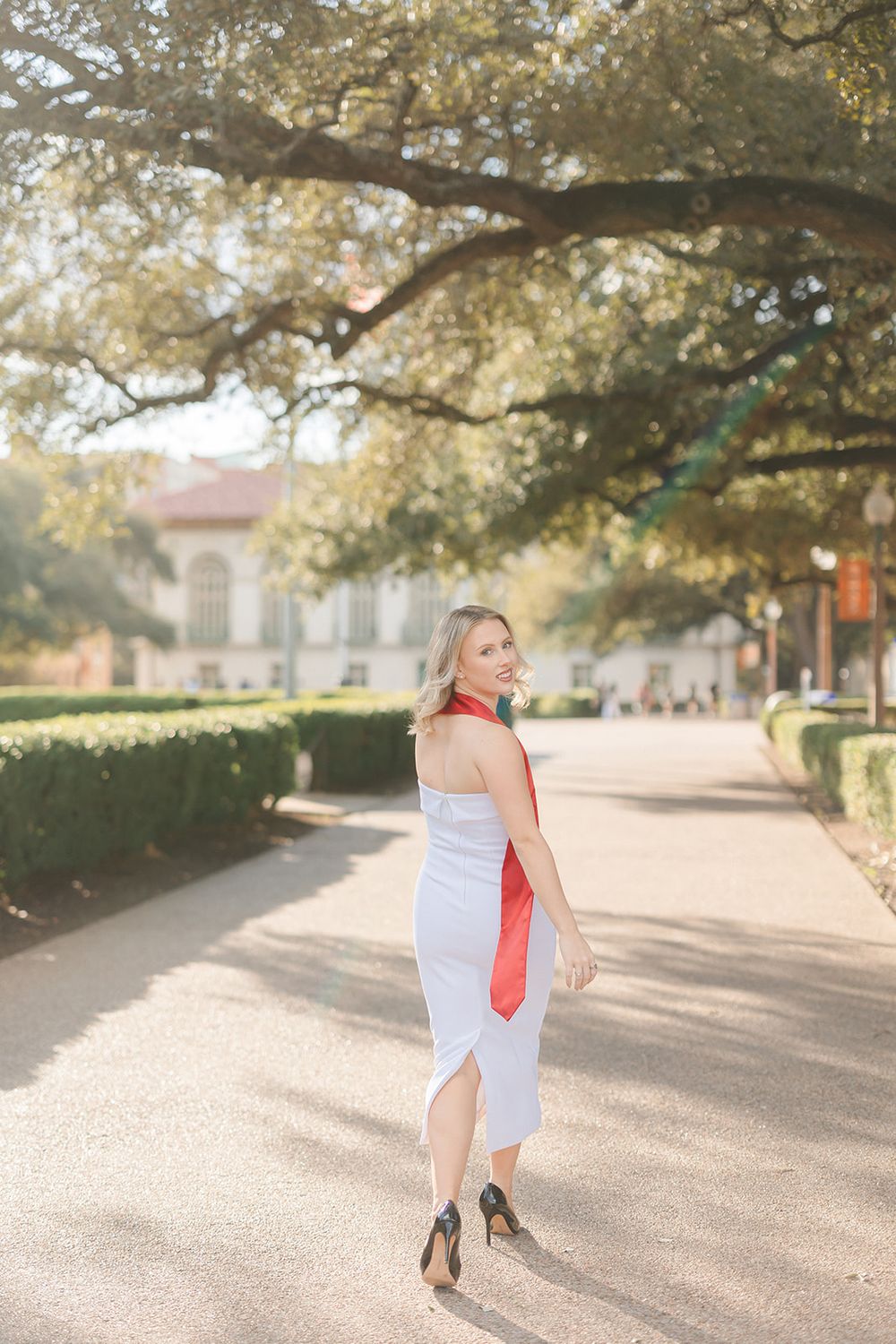 Person in business attire and red scarf walking confidently on a tree-lined path on an autumn day.