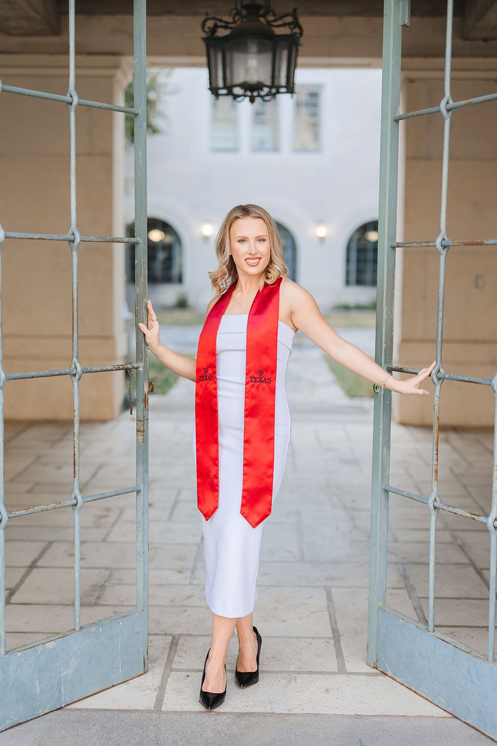 Graduate in white dress and red sash poses by glass doors at University of Texas campus.