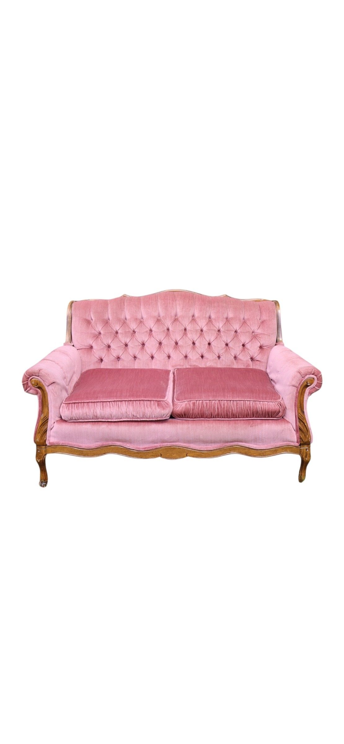 A vintage pink velvet tufted loveseat with wooden frame sits on grass in a wooded outdoor setting.
