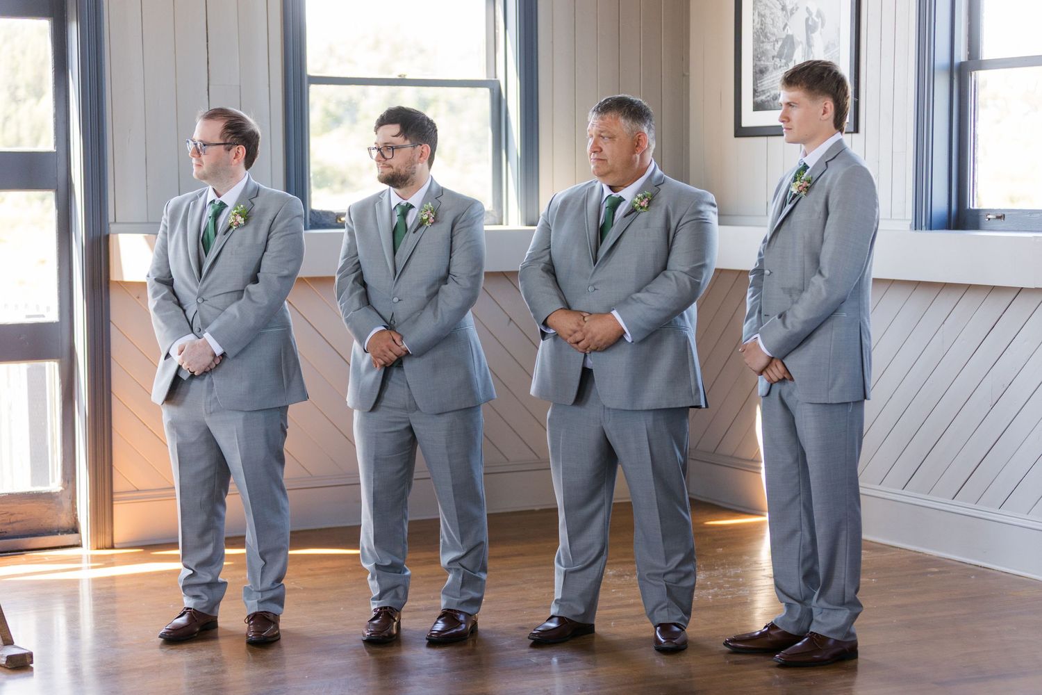 Members of a wedding party in light gray suits stand in profile against white paneled walls during a formal event.